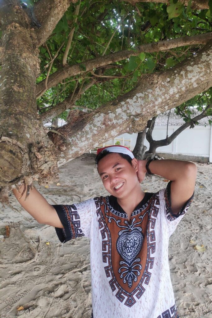 Reymar, WordPress web designer, smiling and posing under a tree on a sandy beach in a patterned shirt.