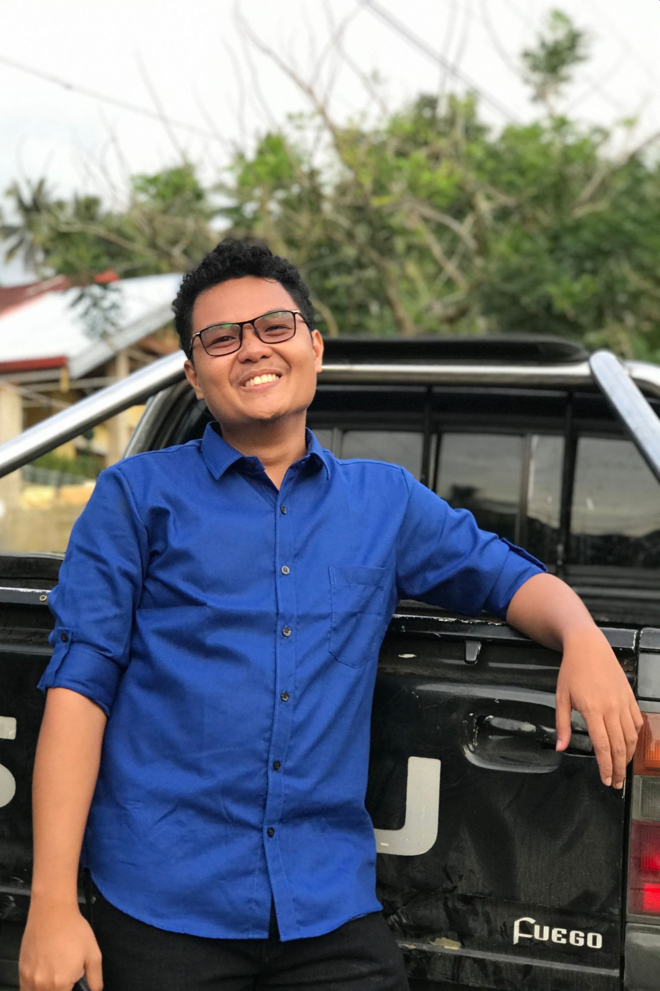 Reymar Pabito smiling while leaning on a black pickup truck, wearing a blue button-up shirt, with trees and houses in the background.